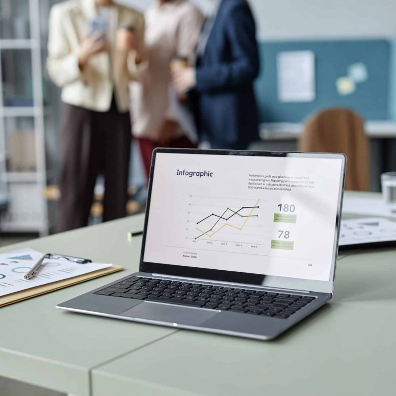 Medium shot with focus on opened laptop displaying business report infographic, water glasses and clipboards on office table, unrecognizable company employees standing in blurred background